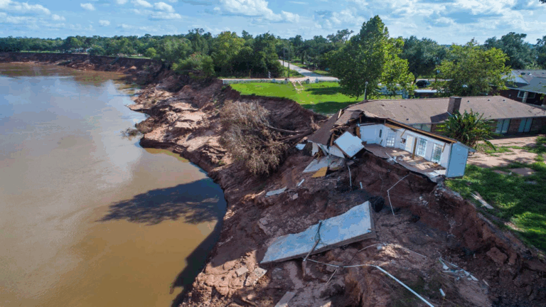Aerial view of severe erosion along the Brazos River in Simonton, Texas, showing a partially collapsed home undermined by the retreating riverbank.