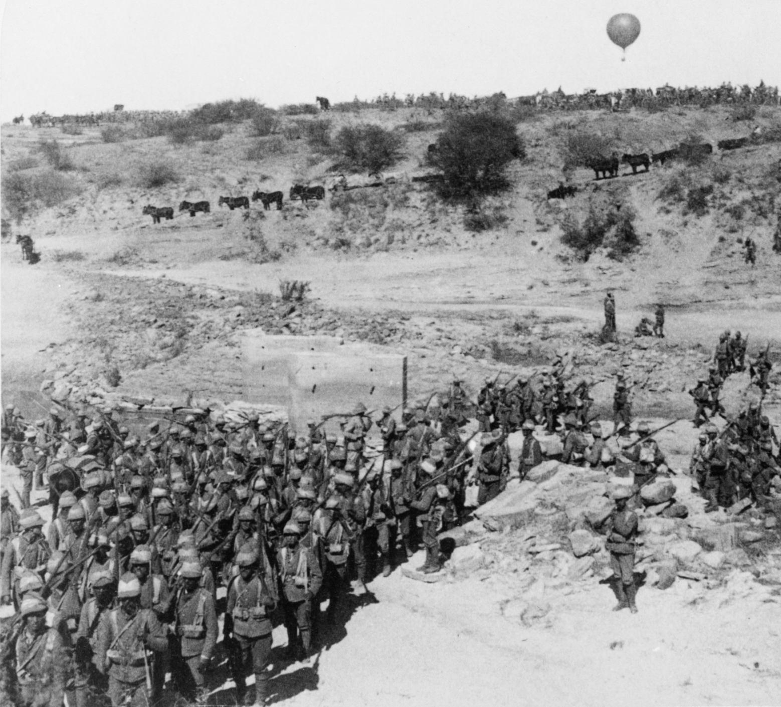 British troops advancing across rocky terrain during the Second Boer War, with a tethered observation balloon visible in the distance.