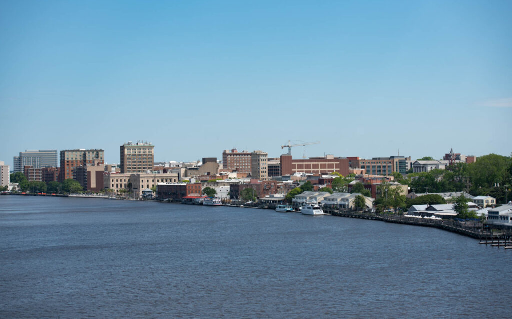 Panoramic view of downtown Wilmington, North Carolina, along the Cape Fear River, showing the waterfront, historic buildings, and modern commercial structures.