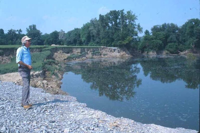 A man stands on the gravel edge of a water-filled sinkhole near Cuylerville, New York, with exposed banks and trees beyond.